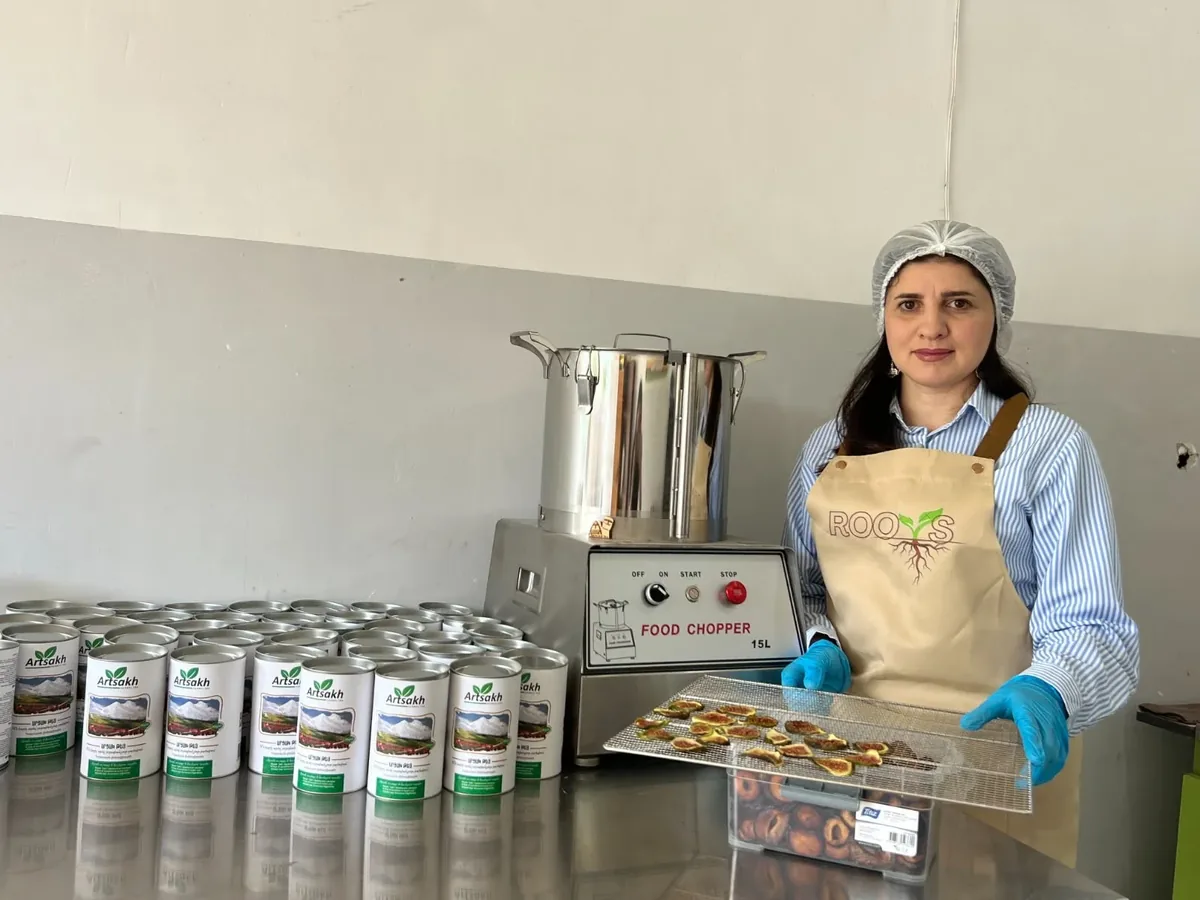 Armine, displaced from Artsakh, holding a tray of dried figs next to her herbal tea tins and food chopper in Vanadzor, Armenia